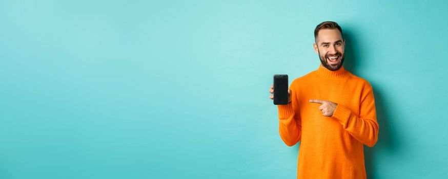 Handsome bearded man in orange sweater, pointing finger at mobile phone screen, showing application smartphone, standing over light blue background.