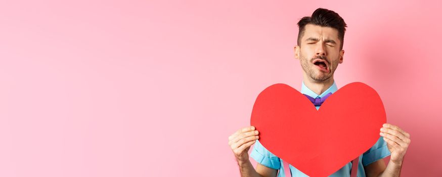 Valentines day concept. Sad and lonely man feeling heartbroken, being rejected, showing big red heart cutout and crying from break-up, standing on pink background.