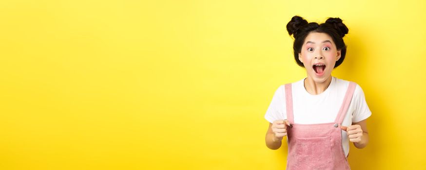 Excited asian woman open mouth, looking fascinated and amazed, receive good news, standing in summer clothes on yellow background.