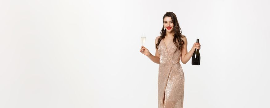 Christmas party and celebration concept. Full length of elegant woman with red lips, luxury dress, holding glass of champagne and bottle, white background.