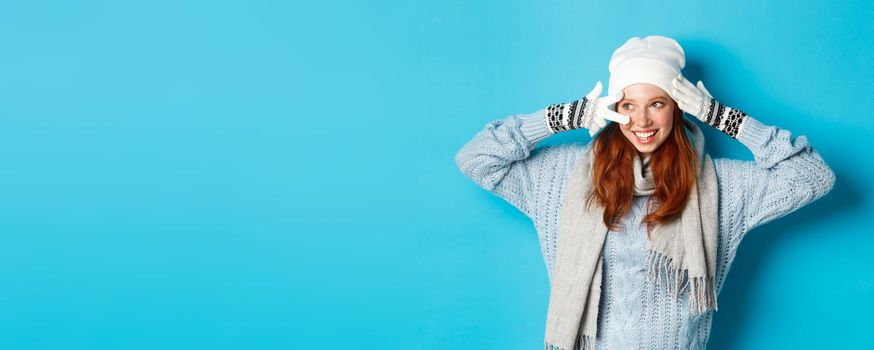 Winter and holidays concept. Cute redhead teen girl in beania, gloves and sweater showing peace sign, looking left at copy space on blue background.