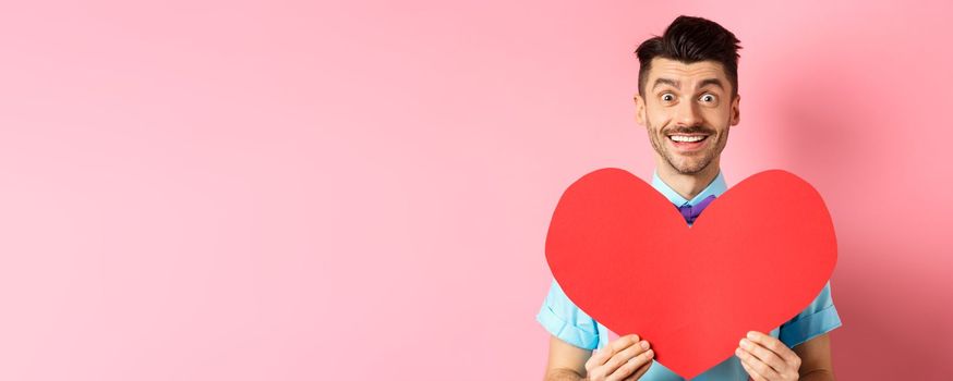 Hopeful man in love showing red heart sign, smiling at camera, waiting for soulmate on Valentines day, standing on pink background.