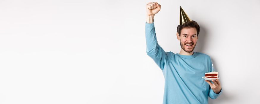Celebration and holidays concept. Happy man celebrating birthday in party hat, holding bday cake and raising hand up in triumph, standing over white background.