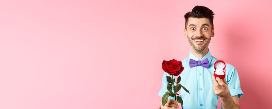 Valentines day. Smiling handsome man asking to marry him, showing engagement ring and red rose, standing romantic on pink background.