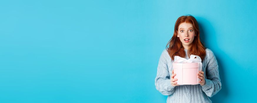 Surprised redhead girl receiving valentines gift, holding box with present and staring at camera amazed, wearing sweater, standing over blue background.