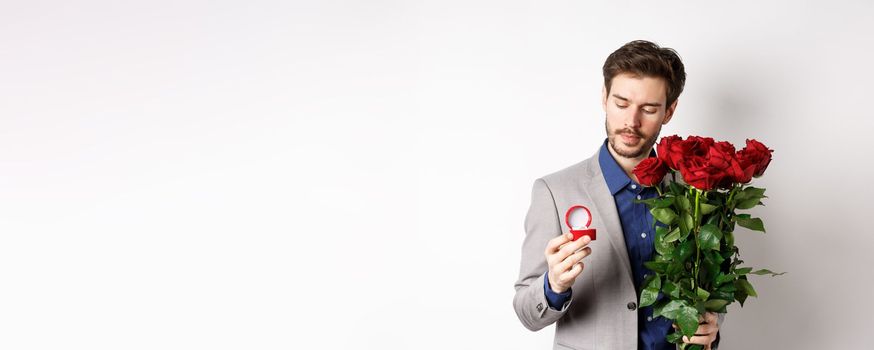 Romantic man in suit looking pensive at engagement ring, going to make a marriage proposal on Valentines day, holding bouquet of roses, standing over white background.