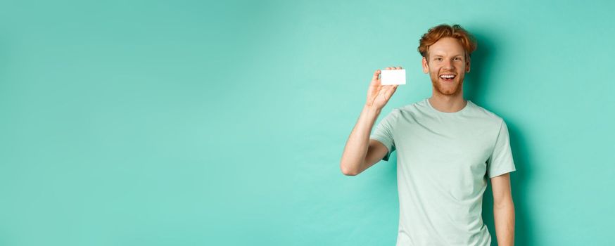 Shopping concept. Cheerful young man in t-shirt showing plastic credit card and smiling, standing over mint background.