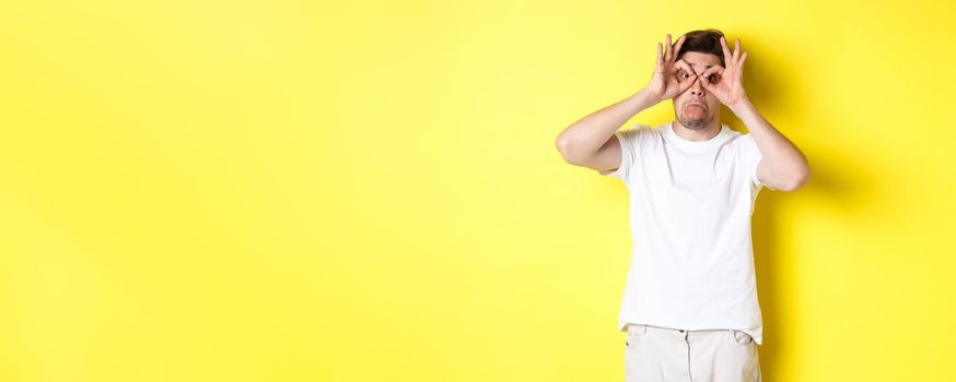 Young man making funny faces and showing tongue, fool around, standing in white t-shirt against yellow background. Copy space
