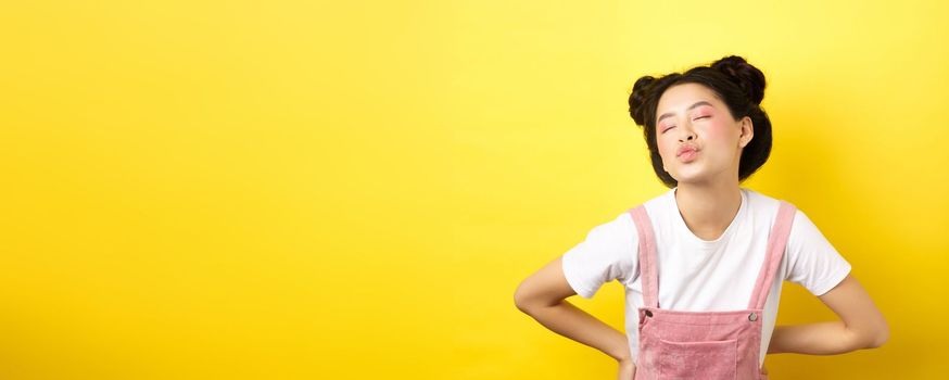 Romantic korean girl close eyes and pucker lips, waiting for sweet kiss on Valentines day, kissing lover, standing on yellow background.