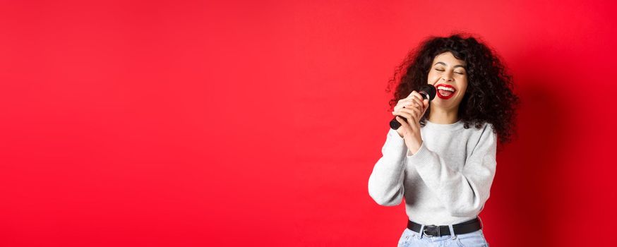 Hobbies and leisure concept. Happy woman singing song in microphone, having fun at karaoke with mic, standing on red background.