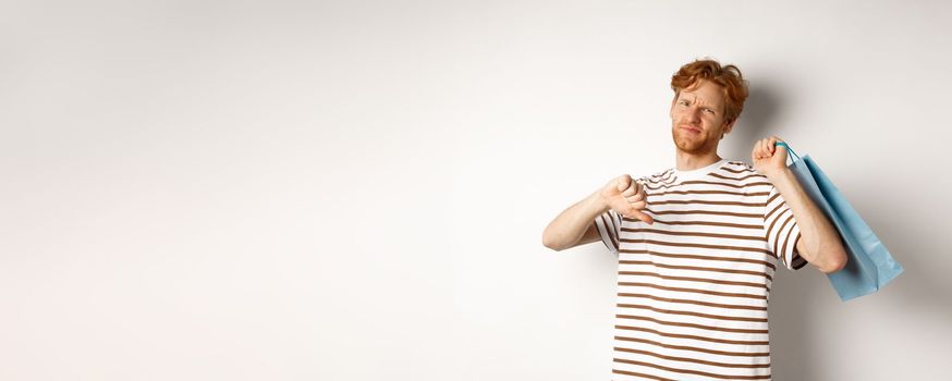 Disappointed young man with red hair and beard showing thumbs-down after bad shopping experience, holding bag over shoulder and frowning upset, white background.