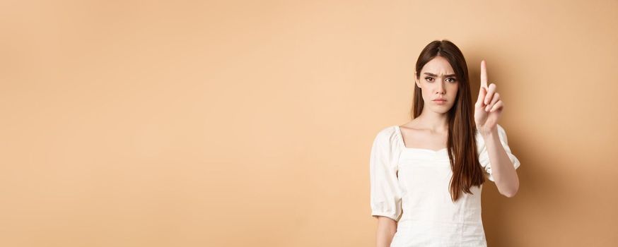 Rule number one. Serious frowning woman showing finger to scold person, look disappointed and angry, standing on beige background.