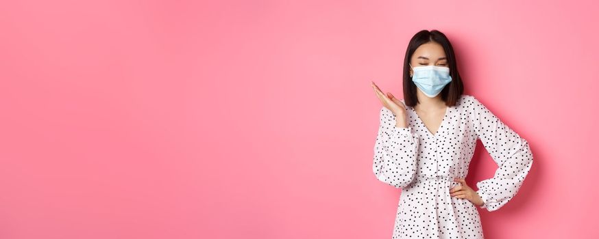 Covid-19, quarantine and lifestyle concept. Beautiful and confident asian woman in dress and face mask looking self-assured, standing sassy against pink background.