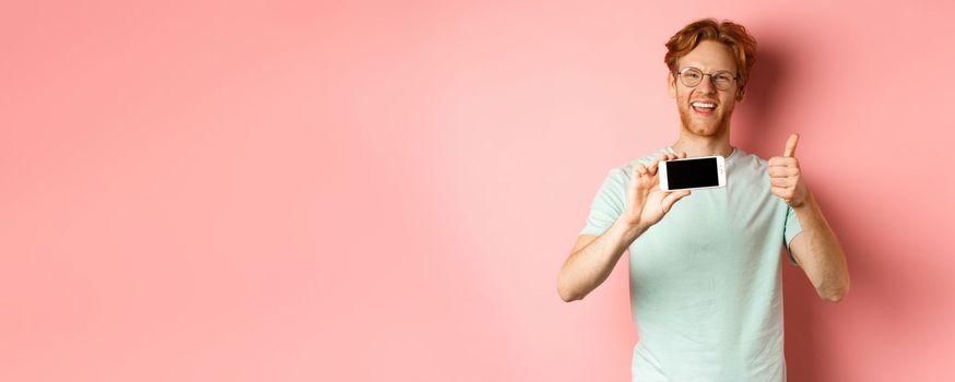 Happy european man with ginger hair and glasses, showing mobile blank screen horizontally and thumbs-up, recommend online promo, pink background.