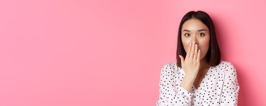 Image of shocked asian girl gossiping, gasping and cover mouth, stare at camera with complete disbelief, standing over pink background.