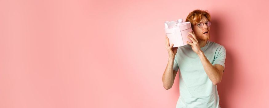 Valentines day and romance concept. Handsome redhead man shaking gift box and wonder what inside, trying to guess present, standing over pink background.