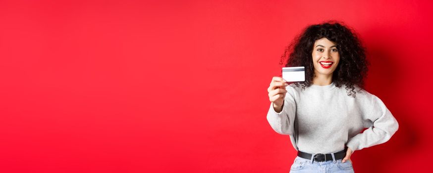 Image of modern woman with curly hair, extending hand and showing plastic credit card, recommending bank or shopping offer, red background.