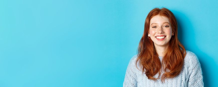 Close-up of cheerful teenage girl with red curly hair, smiling happy at camera, standing against blue background.