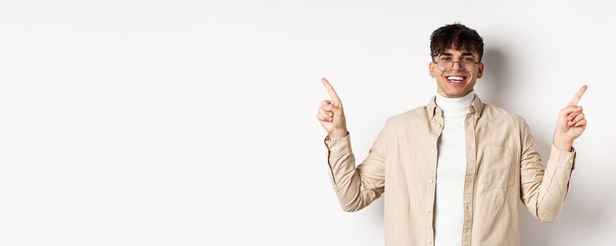 Image of smiling handsome man in glasses pointing fingers sideways, showing advertisements or variants, standing cheerful on white background.