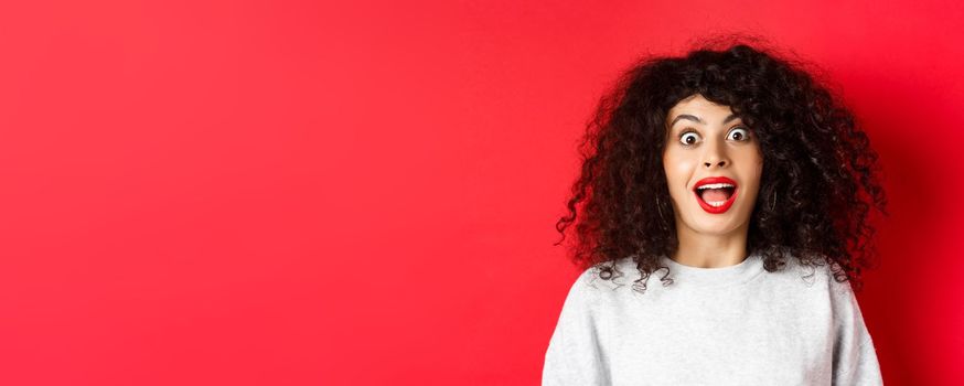 Close-up portrait of excited woman with curly hair, scream surprised and amazed, checking out special deal, standing on red background.