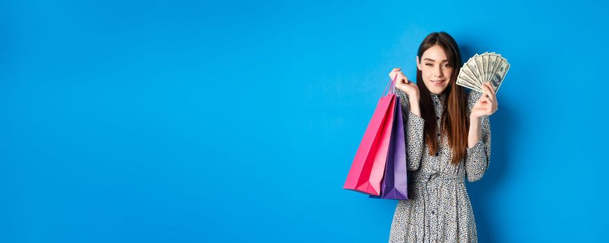 Shopping. Excited attractive woman buying gifts for herself, showing dollar bills and color bags from shop, smiling satisfied, standing on blue background.