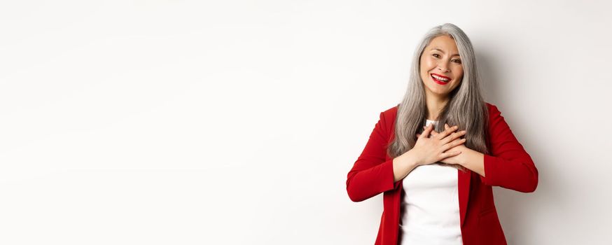 Business concept. Mature asian woman with red lips and blazer, holding hands on heart and smiling thankful, looking grateful at camera, standing over white background.