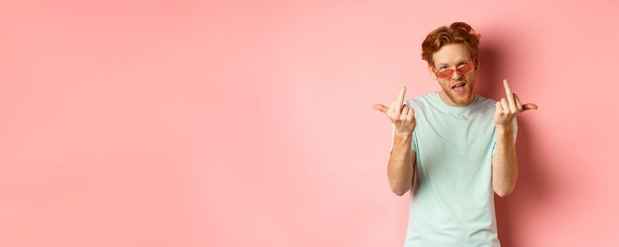 Arrogant and careless redhead man in sunglasses dont give a fuck, showing middle fingers at camera and frowning, standing over pink background.