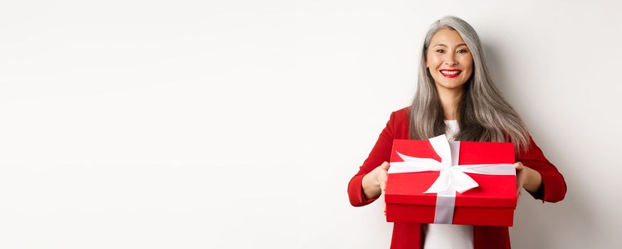 Elegant senior woman giving you present. Asian lady holding red gift box and smiling, standing over white background.