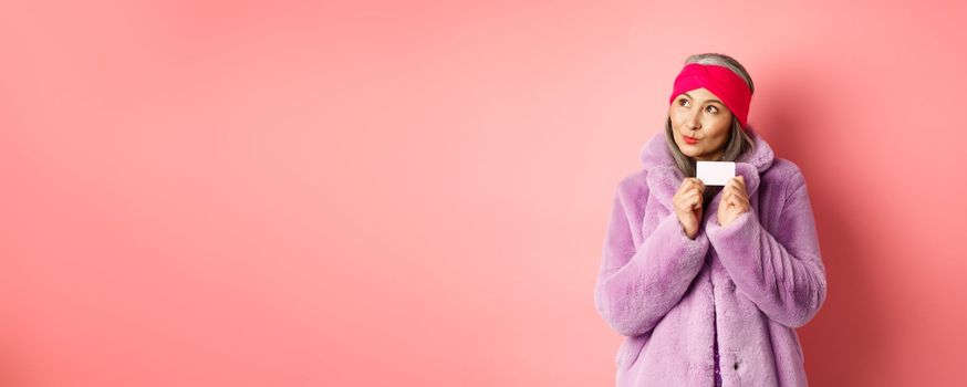 Shopping and fashion concept. Fashionable asian senior woman thinking about buying gifts, holding credit card and looking thoughtful at upper left corner, pink background.