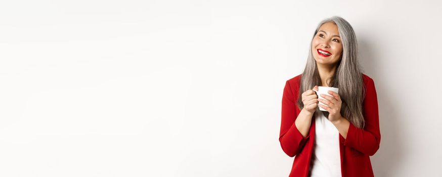 Business people concept. Smiling asian businesswoman having coffee break, holding warm mug and looking upper left corner dreamy, white background.