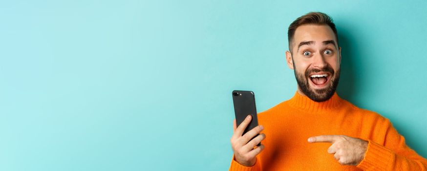 Close-up of impressed man showing something on mobile phone, pointing at smartphone and gasping amazed, standing in orange sweater over light blue background.