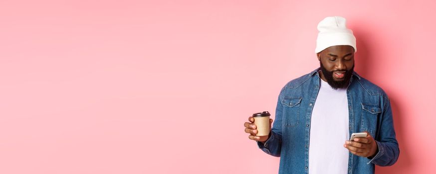 Image of stylish Black man hipster drinking takeaway coffee, reading message on phone, standing over pink background.