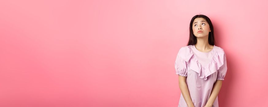 Dreamy and sad asian woman look at upper left corner logo, standing pensive and gloomy in pink romantic dress, white background.