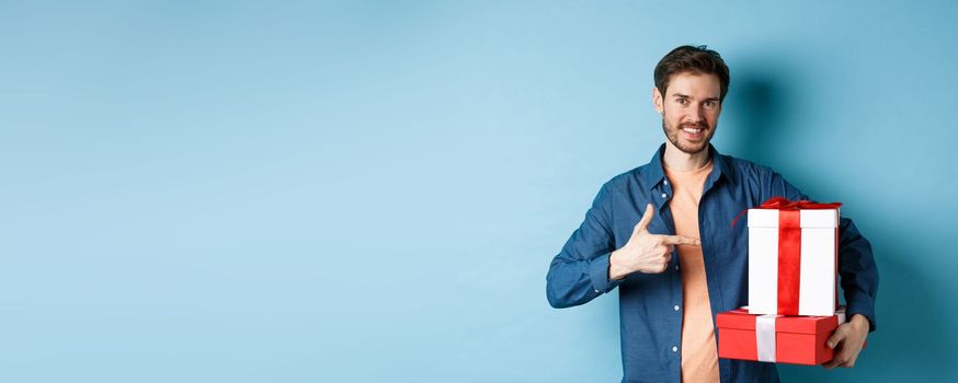 Attractive boyfriend pointing finger at valentines day surprise gifts, showing romantic presents for girlfriend, standing against blue background.