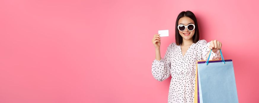 Beautiful asian woman in sunglasses going shopping, holding bags and showing credit card, standing over pink background.