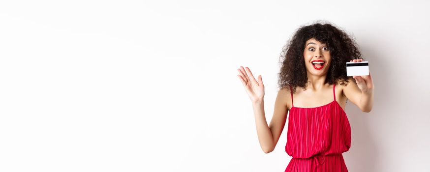 Shopping. Amazed curly-haired woman in red dress, showing plastic credit card and scream from excitement, standing on white background.