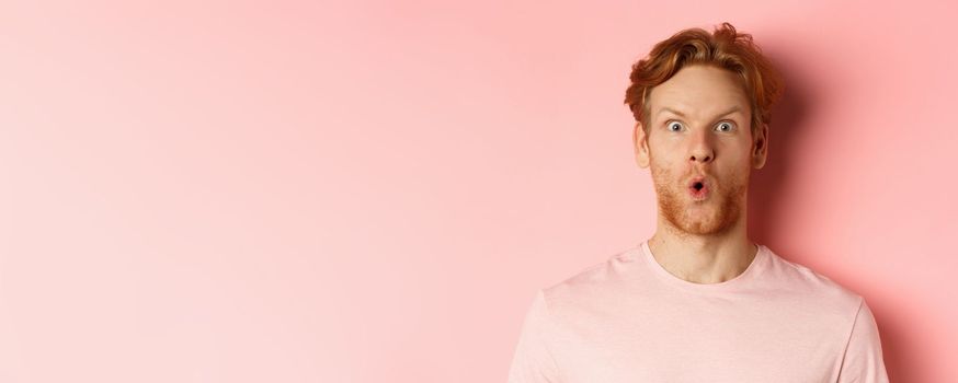 Headshot of young redhead man with beard, saying wow and staring at camera amazed, checking out promo deal, standing over pink background.