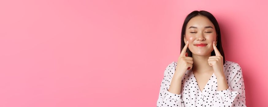 Beauty and lifestyle concept. Close-up of beautiful asian woman poking cheeks with closed eyes, smiling satisfied, standing over pink background.