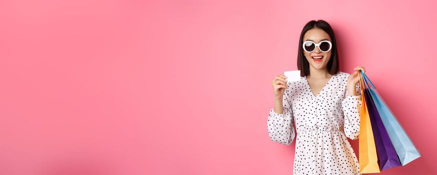 Beautiful asian woman in sunglasses going shopping, holding bags and showing credit card, standing over pink background.