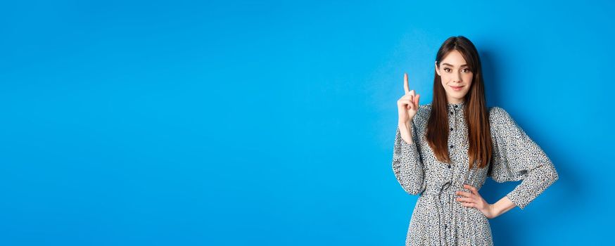 Young beautiful lady in dress with natural long hair, showing number one with finger and smiling, standing against blue background.