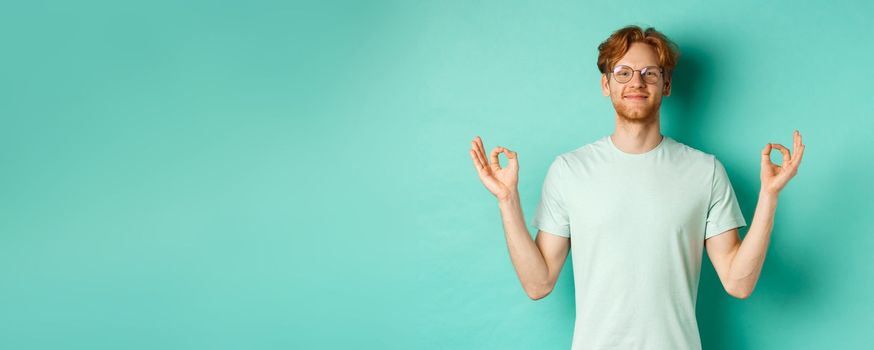 Calm and relaxed redhead man in glasses smiling, holding hands spread sideways with om mudra gesture, meditating peaceful, standing over mint background.