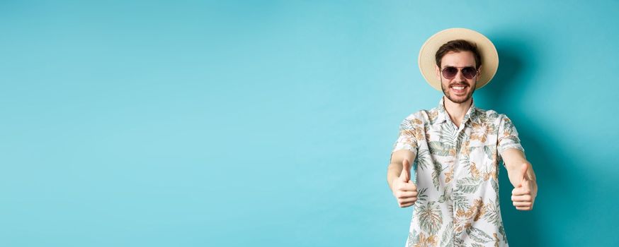 Happy tourist in sunglasses and straw hat showing thumbs up and smiling, enjoying summer holiday, standing on white background.