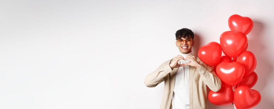 Valentines day and romance concept. Handsome young man standing near red hearts balloons and showing heart gesture, being in love and smiling, white background.