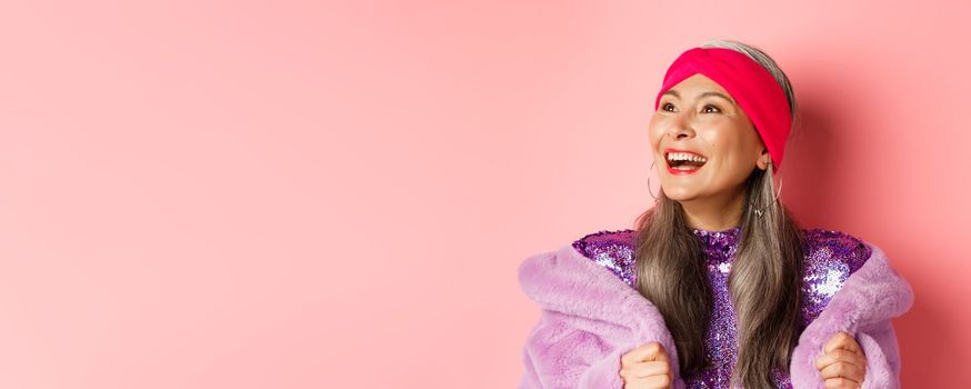 Fashion. Close-up of happy asian senior woman smiling, looking left with cheerful face, standing in purple faux fur coat, pink background.