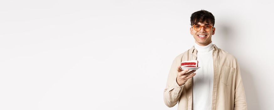 Happy caucasian guy celebrating birthday, holding cake with candle and smiling cheerful at camera, standing on white background.