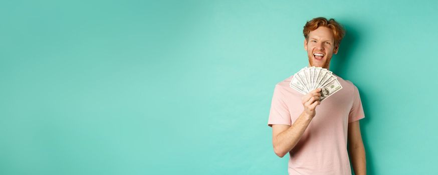 Happy redhead man in t-shirt showing money in dollars and smiling, making smug faces after winning cash, standing over turquoise background.