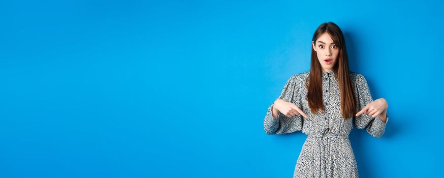 Excited young woman in dress pointing fingers down at logo, looking intrigued at camera, standing on blue background.