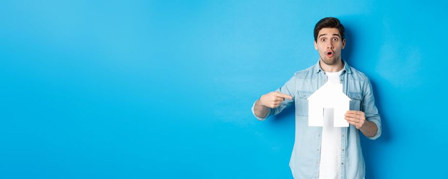 Insurance, mortgage and real estate concept. Surprised young man pointing at house card model and looking at camera, standing against blue background.