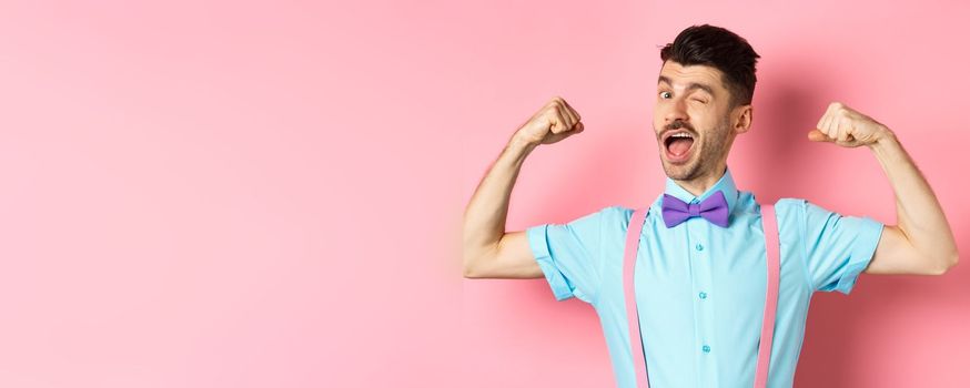 Strong and funny guy with french moustache, flexing biceps and winking at camera, show-off his strengths, standing over pink background.