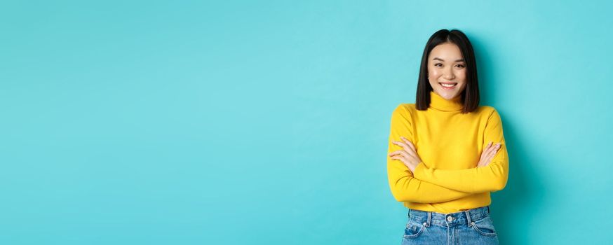 Confident and stylish asian woman cross arms on chest and smiling, standing over blue background.